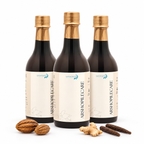Three dark glass bottles of Sanadana Ayurvedic Arshopilecare oil with gold caps, arranged on a white background with whole spices and nuts in front.