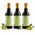 Three dark glass bottles of DHATRYARISHTAM with gold caps and light green labels, arranged on a white background with whole and sliced green amla fruits around the base.