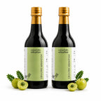 Two dark glass bottles of Dhiatrayishtan with gold caps and light green labels, arranged side by side on a white background with whole and sliced green amla and leaves at their bases.